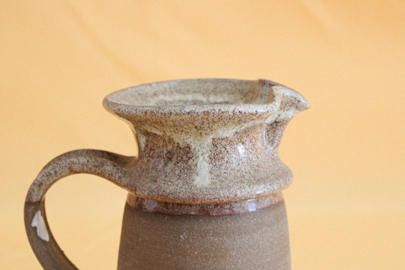 May include: A brown and white ceramic pitcher with a textured glaze. The pitcher has a wide, round spout and a handle.