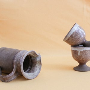May include: Two brown ceramic cups with white glaze stacked on top of each other. The cups have a footed base. A brown ceramic pitcher with white glaze is lying on its side.
