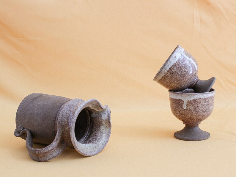 May include: Two brown ceramic cups with white glaze stacked on top of each other. The cups have a footed base. A brown ceramic pitcher with white glaze is lying on its side.