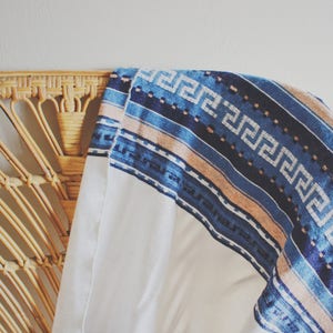 May include: A white throw blanket with a blue and tan patterned border. The blanket is draped over a wicker chair.
