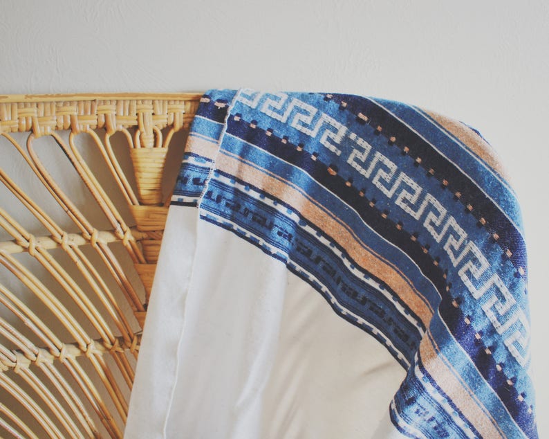 May include: A white throw blanket with a blue and tan patterned border. The blanket is draped over a wicker chair.