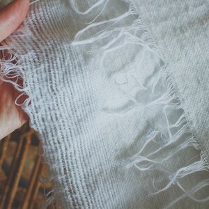 May include: A white fabric with a frayed edge. The fabric has a woven texture and is slightly wrinkled.