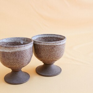 May include: Two brown ceramic cups with a white rim and a speckled glaze. The cups are on a light orange surface.