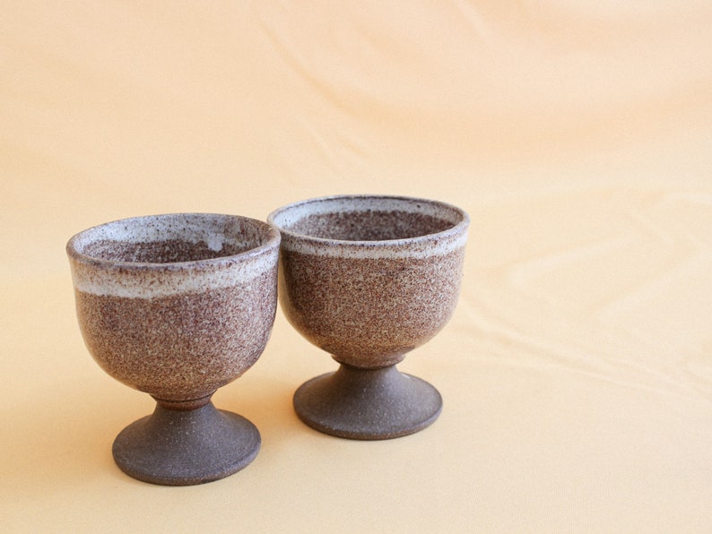 May include: Two brown ceramic cups with a white rim and a speckled glaze. The cups are on a light orange surface.