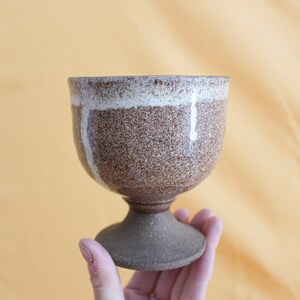May include: A brown and white ceramic goblet with a speckled glaze. The goblet has a wide base and a rounded bowl.