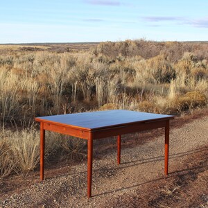 Puede incluir: Una mesa de comedor rectangular de madera oscura con una extensi&oacute;n de hoja abatible. La mesa est&aacute; sentada en un camino de grava en un campo de hierba seca.
