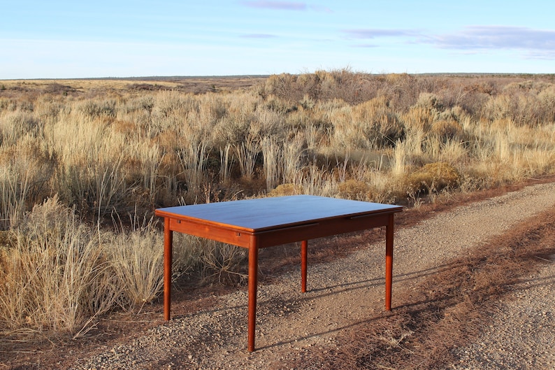 Puede incluir: Una mesa de comedor rectangular de madera oscura con una extensi&oacute;n de hoja abatible. La mesa est&aacute; sentada en un camino de grava en un campo de hierba seca.