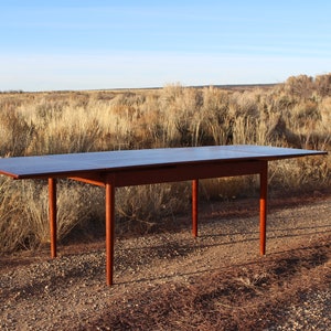 Puede incluir: Una mesa de comedor extensible de madera oscura con una hoja, de pie en un camino de grava frente a un campo de matorrales secos.