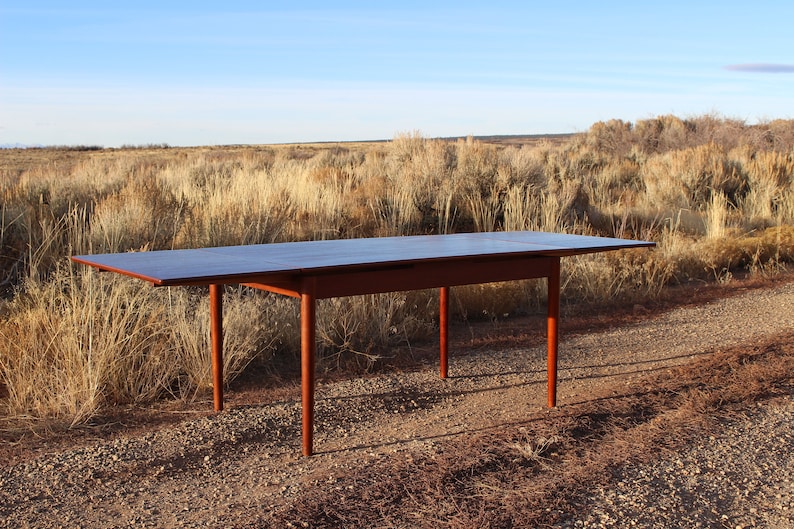 Puede incluir: Una mesa de comedor extensible de madera oscura con una hoja, de pie en un camino de grava frente a un campo de matorrales secos.