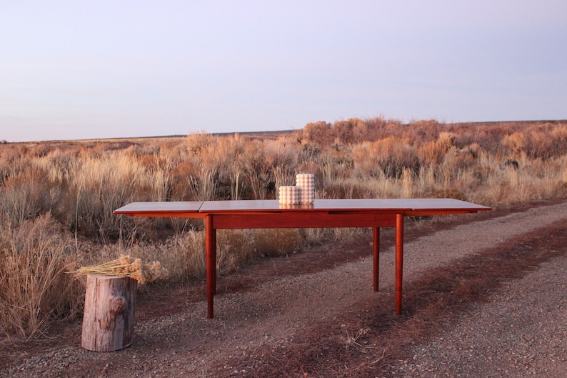 Puede incluir: Una mesa de comedor extensible de madera oscura con un acabado de madera natural. La mesa est&aacute; instalada al aire libre en un camino de grava con un paisaje seco y herboso en el fondo. Dos contenedores cil&iacute;ndricos con patrones blancos y marrones est&aacute;n sobre la mesa.