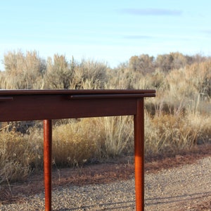 Puede incluir: Una mesa de comedor de madera oscura con dos hojas, que extienden la longitud de la mesa. La mesa est&aacute; de pie en un camino de grava con matorrales secos en el fondo.