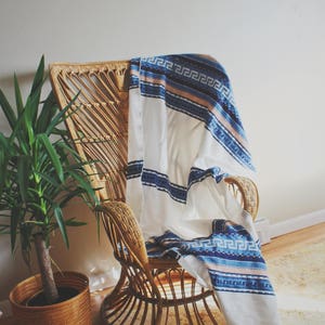 May include: A wicker chair with a white and blue patterned throw blanket draped over it. The chair is sitting on a rug with a light brown and white pattern. A potted plant is in the foreground.