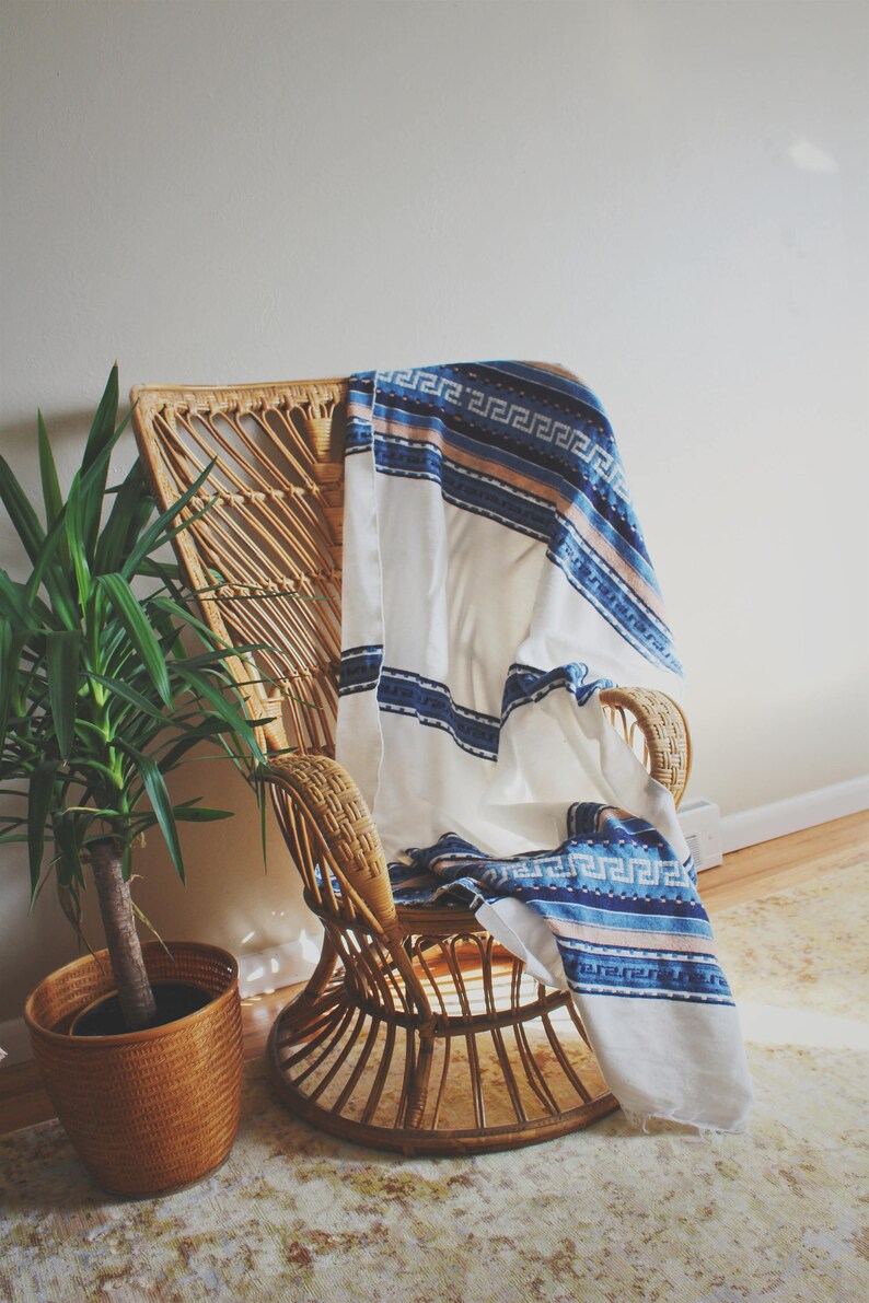 May include: A wicker chair with a white and blue patterned throw blanket draped over it. The chair is sitting on a rug with a light brown and white pattern. A potted plant is in the foreground.