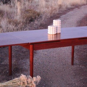 Puede incluir: Una mesa de comedor extensible de madera oscura con dos jarrones de cer&aacute;mica blanca sobre la superficie. La mesa est&aacute; colocada en un camino de grava con matorrales secos en el fondo.