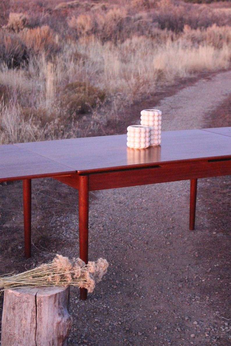 Puede incluir: Una mesa de comedor extensible de madera oscura con dos jarrones de cer&aacute;mica blanca sobre la superficie. La mesa est&aacute; colocada en un camino de grava con matorrales secos en el fondo.