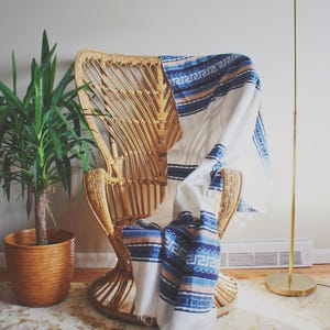 May include: A white throw blanket with blue and pink stripes and a geometric pattern draped over a wicker chair. A potted plant and a gold floor lamp are in the background.