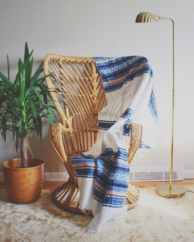 May include: A white throw blanket with blue and pink stripes and a geometric pattern draped over a wicker chair. A potted plant and a gold floor lamp are in the background.