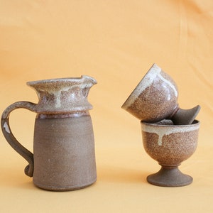 May include: A brown ceramic pitcher with a white glaze and two brown ceramic cups with white glaze. The cups are stacked on top of each other.