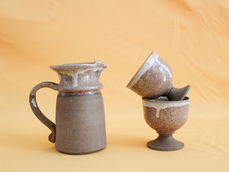May include: A brown ceramic pitcher with a white glaze and two brown ceramic cups with white glaze. The cups are stacked on top of each other.
