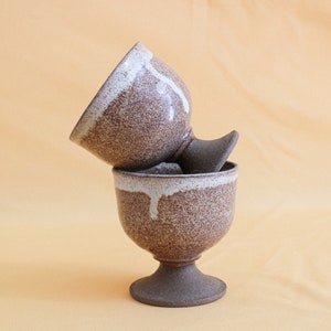 May include: Two brown ceramic cups with white glaze, stacked on top of each other. The cups have a speckled brown glaze and a white glaze that drips down the sides. The cups have a pedestal base.