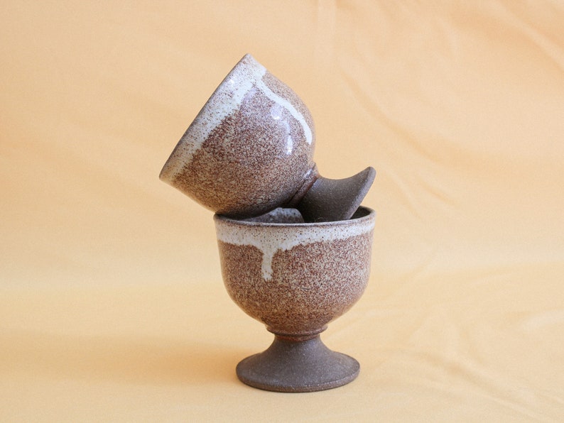 May include: Two brown ceramic cups with white glaze, stacked on top of each other. The cups have a speckled brown glaze and a white glaze that drips down the sides. The cups have a pedestal base.