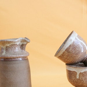 May include: A brown and white ceramic pitcher with a wide mouth and a handle. Next to it is a brown and white ceramic cup with a white glaze on the inside. The cup is sitting on top of another brown and white ceramic cup.