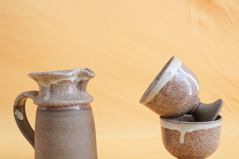 May include: A brown and white ceramic pitcher with a wide mouth and a handle. Next to it is a brown and white ceramic cup with a white glaze on the inside. The cup is sitting on top of another brown and white ceramic cup.