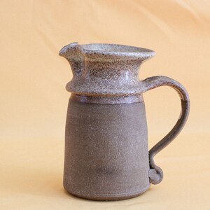 May include: A brown and white ceramic pitcher with a speckled glaze. The pitcher has a wide mouth and a handle that curves up and out.