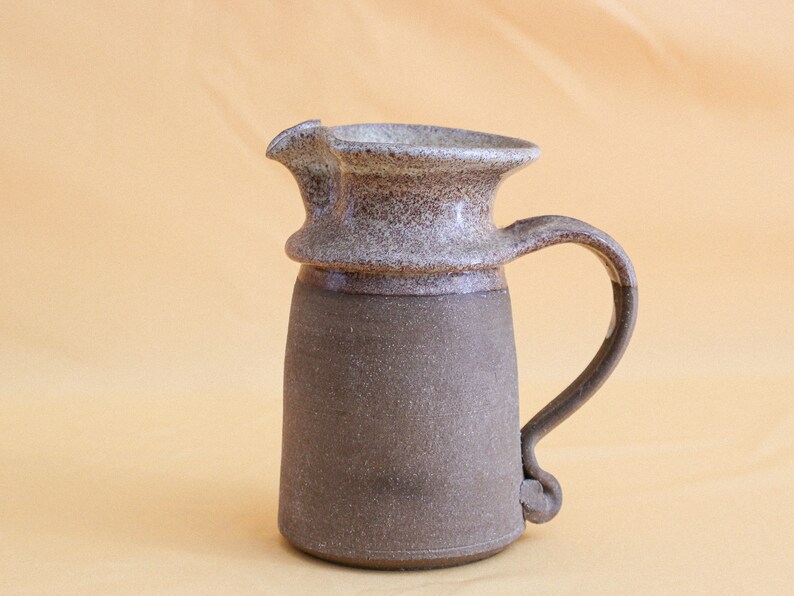May include: A brown and white ceramic pitcher with a speckled glaze. The pitcher has a wide mouth and a handle that curves up and out.