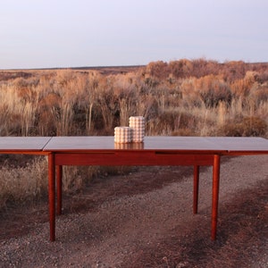 Puede incluir: Una mesa de comedor extensible de madera oscura con dos macetas de cer&aacute;mica blanca sobre la superficie. La mesa est&aacute; situada sobre un fondo de matorrales secos y marrones.