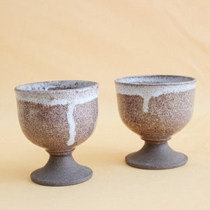 May include: Two brown and white ceramic cups with a speckled glaze and a white drip design. The cups have a pedestal base.