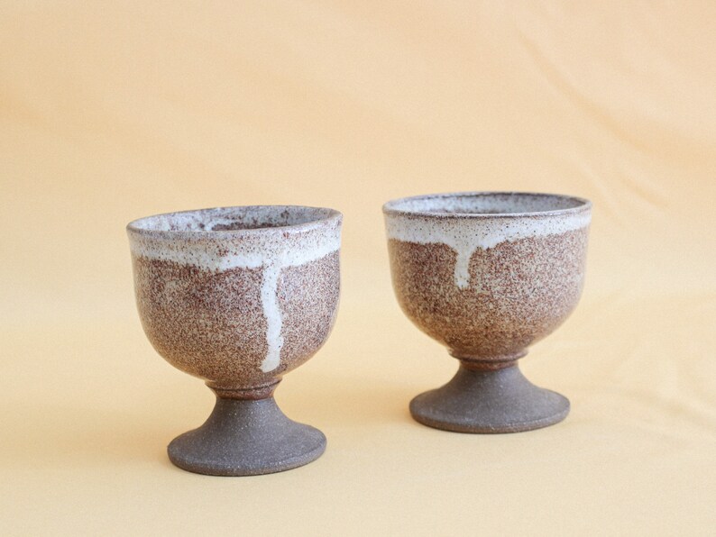 May include: Two brown and white ceramic cups with a speckled glaze and a white drip design. The cups have a pedestal base.