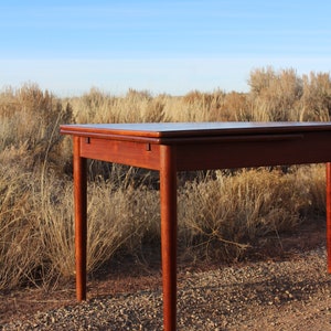 Puede incluir: Una mesa de madera oscura con una tapa rectangular y cuatro patas. La mesa est&aacute; sentada en una superficie de grava con hierba seca en el fondo.