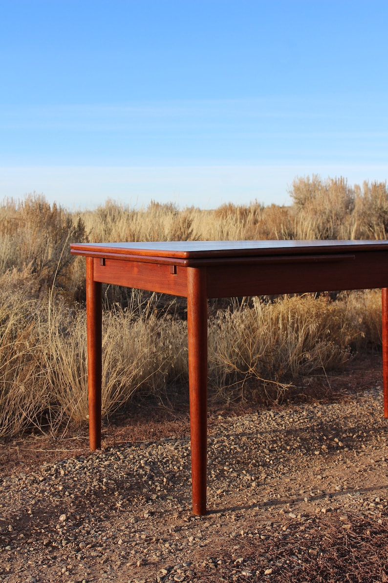 Puede incluir: Una mesa de madera oscura con una tapa rectangular y cuatro patas. La mesa est&aacute; sentada en una superficie de grava con hierba seca en el fondo.