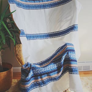 May include: A white throw blanket with blue and pink stripes and a geometric pattern. The blanket is draped over a wicker chair.