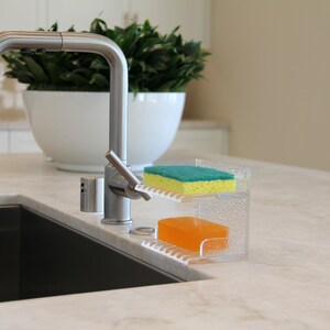 May include: A clear plastic dish soap and sponge holder with a green sponge and a yellow bar of soap. The holder is sitting on a white countertop next to a stainless steel kitchen sink.