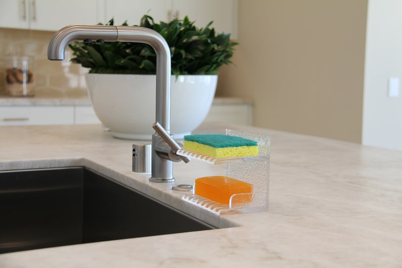 May include: A clear plastic dish soap and sponge holder with a green sponge and a yellow bar of soap. The holder is sitting on a white countertop next to a stainless steel kitchen sink.