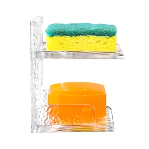 May include: A clear plastic dish soap and sponge holder with two tiers. The top tier holds a green and yellow sponge. The bottom tier holds two orange bars of soap.