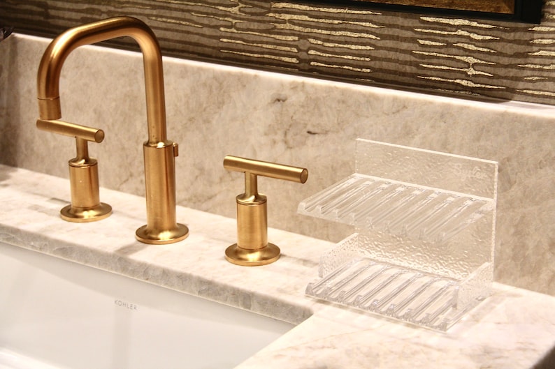 May include: Gold bathroom faucet with a single lever handle and a clear plastic soap dish with two compartments. The faucet is labeled "Kohler".
