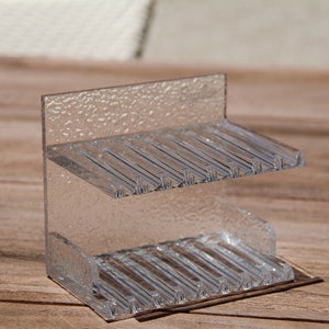 May include: A clear plastic two-tiered stand with a textured surface. The stand has multiple slots on each tier.