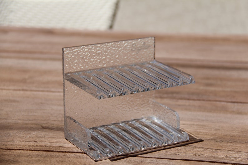 May include: A clear plastic two-tiered stand with a textured surface. The stand has multiple slots on each tier.