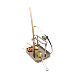 May include: A grey metal kitchen utensil holder with a glass lid. The holder has a rack for spoons and a stand for a wooden spatula. There are three spoons with food residue on the bottom of the holder.
