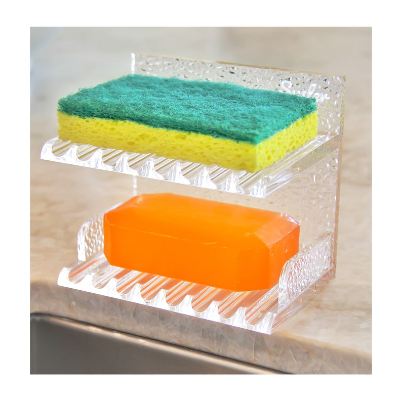 May include: A clear plastic dish soap and sponge holder with two tiers. The top tier holds a green and yellow sponge and the bottom tier holds a rectangular orange bar of soap.
