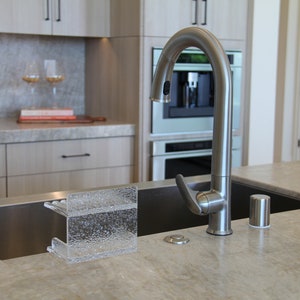 May include: A stainless steel kitchen sink with a modern faucet and a clear glass dish rack. The sink is set into a countertop made of a light gray speckled material.