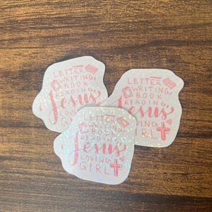 May include: Three stickers with a white background and a holographic effect. Each sticker has the words "Letter Writing, Book Reading, Jesus Loving Girl" in pink script. A cross is also featured.