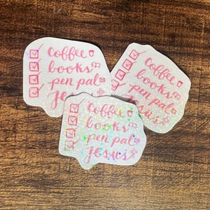 May include: Three holographic stickers with pink text. Each sticker lists "coffee, books, pen pal, Jesus" with check boxes. The stickers have a glittery, iridescent effect, and are on a wooden surface.