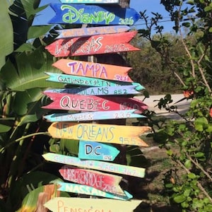May include: A collection of colorful wooden arrow signs pointing in various directions, attached to a tree trunk. Each sign displays a destination name and a distance in miles, including Tahoe, Disney, Las Vegas, and Tampa.