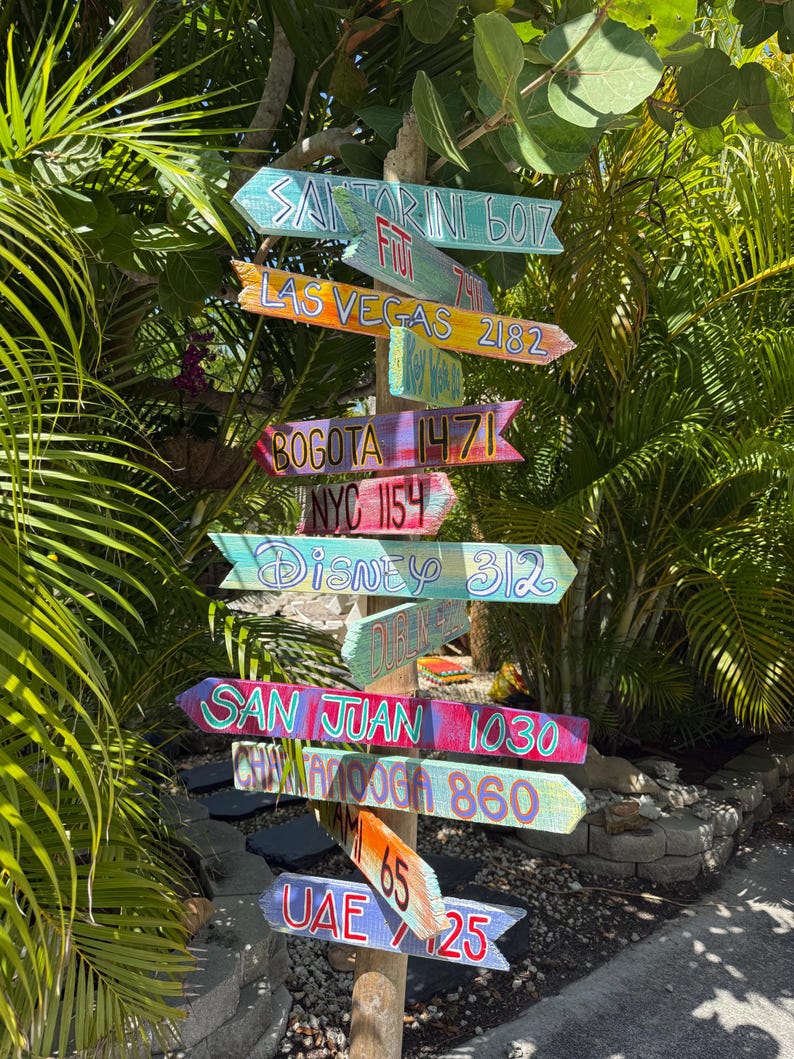 May include: A wooden signpost with multiple colorful directional signs pointing in various directions. The signs display names of cities like Santorini, Las Vegas, Bogota, and Disney, along with distance measurements in miles. The signpost is surrounded by lush green foliage.