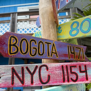 May include: Wooden directional signs with painted text. The signs point in different directions, with destinations including "Bogota 1471" and "NYC 1154". The signs are mounted on a wooden post, with a blue building in the background.