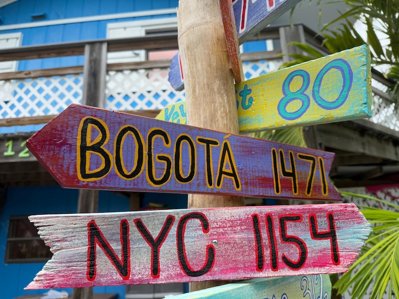 May include: Wooden directional signs with painted text. The signs point in different directions, with destinations including "Bogota 1471" and "NYC 1154". The signs are mounted on a wooden post, with a blue building in the background.